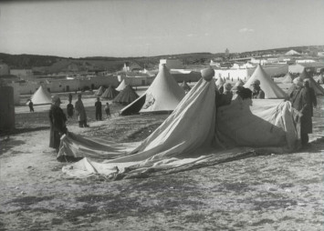 Photo en noir et blanc montrant un camp de réfugiés avec de grandes tentes en train d'être montées