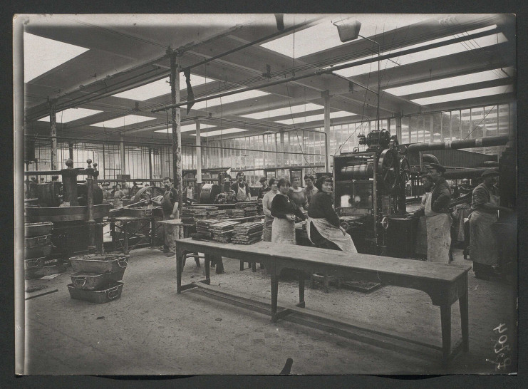 Photo en noir et blanc d'une usine de chocolat, montrant des machines de production, des formes de tablettes de chocolat et des ouvriers à l'arrêt qui regardent l'objectif